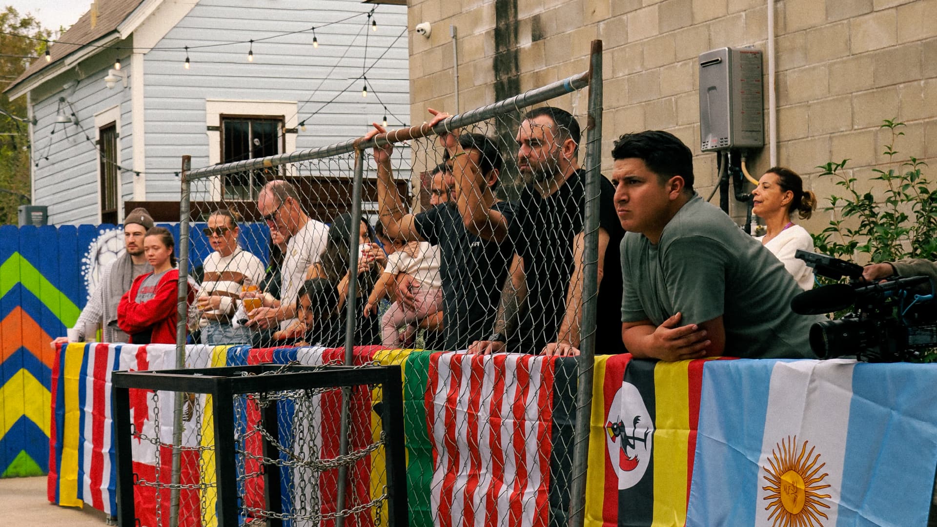 Spectators leaning on the flag-draped fence around the East-side pitch