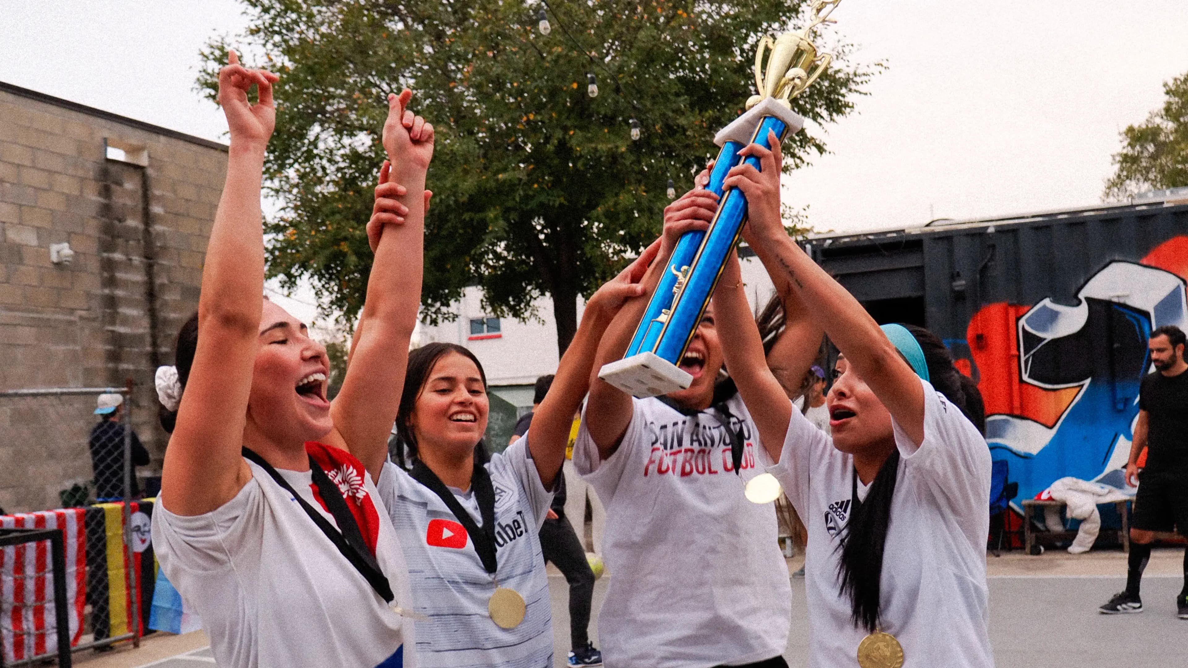 Women’s side lifting the trophy on the East-side court, medals on, crowd behind them