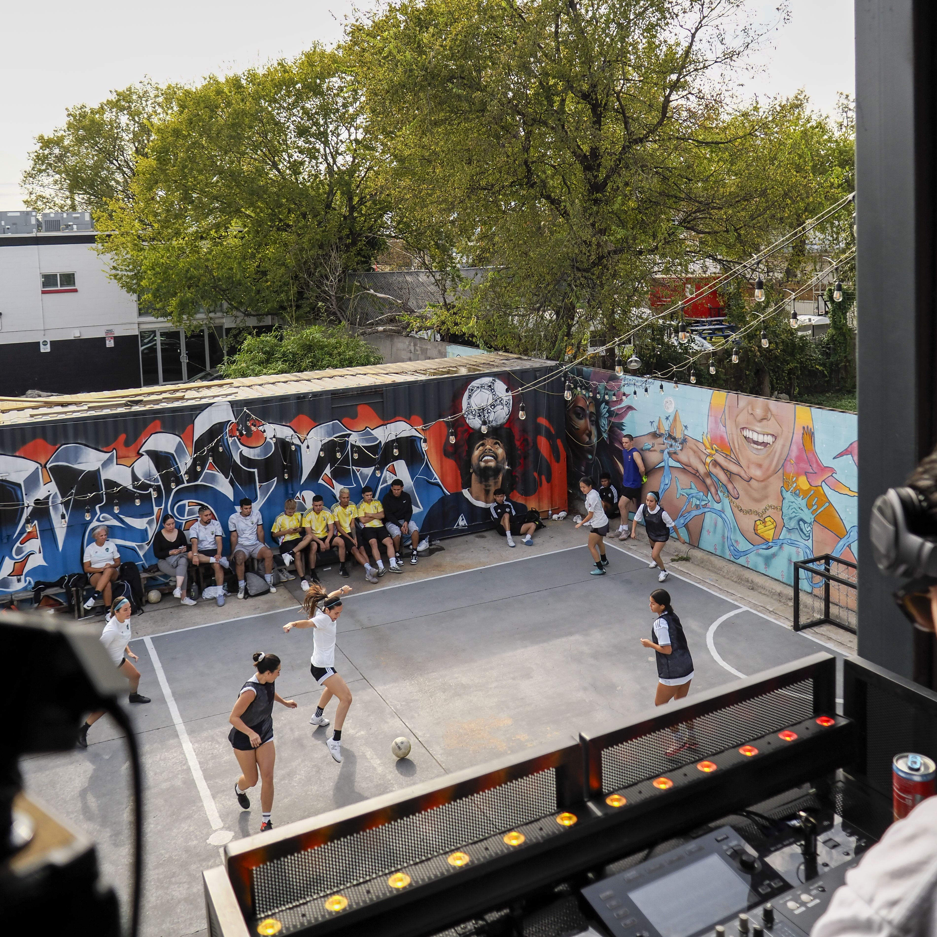 Overhead view of the East-side court in full swing — match on, murals behind, and the DJ booth in the foreground
